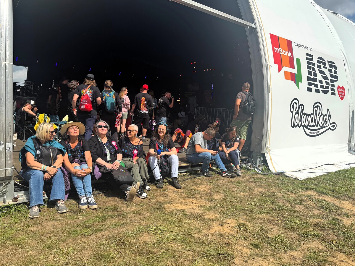 A group of women sit on the grass at the entrance to the ASP tent with Pol'and'Rock and mBank logos. In the background you can see people walking by and the scene inside the tent.